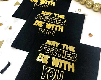 以下が含まれることがあります： 「MAY THE FORTIES BE WITH YOU」という金箔の文字が太字のスタイリッシュなフォントで印刷された、3枚の正方形の黒い紙ナプキン。ナプキンは白い表面に、ずらして重ねた対角線状に配置されています。