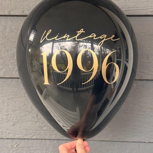 May include: A black balloon with gold text reading "Vintage 1996." The balloon is held by a hand. The balloon reflects the surrounding environment.