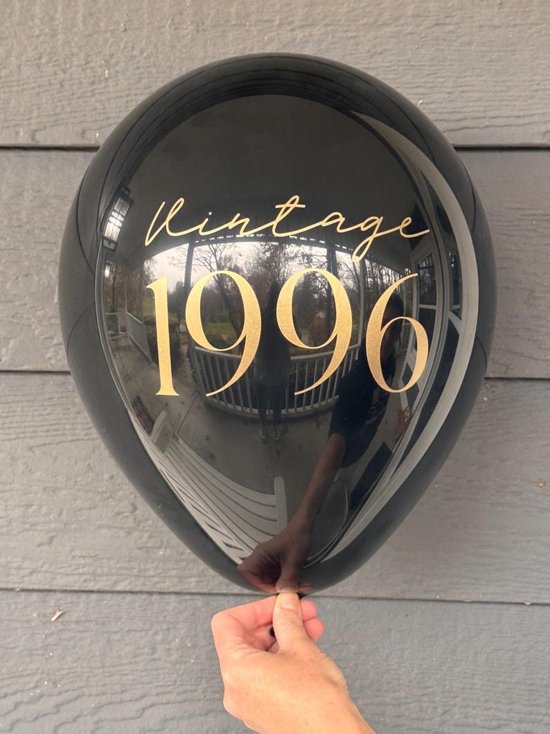 May include: A black balloon with gold text reading "Vintage 1996." The balloon is held by a hand. The balloon reflects the surrounding environment.