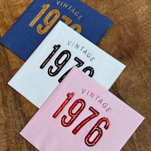 May include: Three square paper napkins in blue, white, and pink. Each napkin has the word "VINTAGE" above the year, with the year 1970 or 1976 in a metallic finish. The napkins are on a wooden surface.