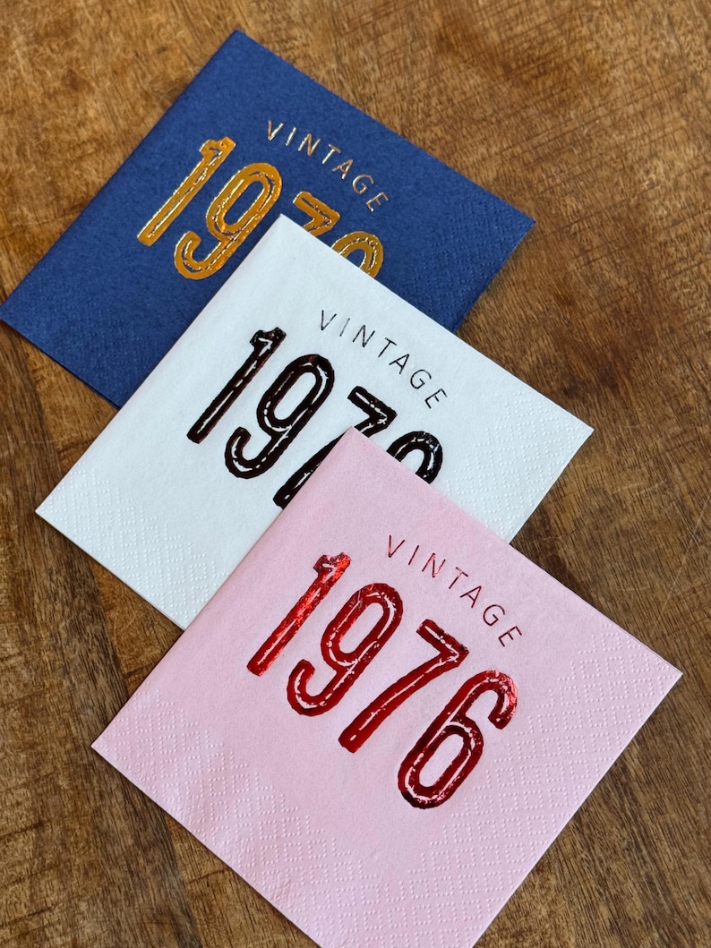 May include: Three square paper napkins in blue, white, and pink. Each napkin has the word "VINTAGE" above the year, with the year 1970 or 1976 in a metallic finish. The napkins are on a wooden surface.
