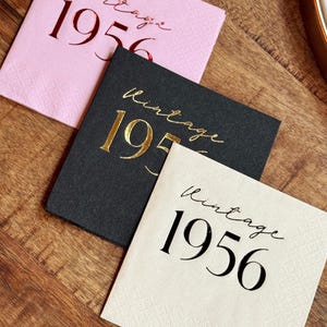May include: Three square paper napkins with the text "Vintage 1956". One napkin is pink with red text, one is black with gold text, and one is off-white with black text. The napkins are on a wooden surface.