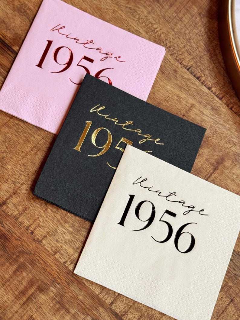 May include: Three square paper napkins with the text "Vintage 1956". One napkin is pink with red text, one is black with gold text, and one is off-white with black text. The napkins are on a wooden surface.