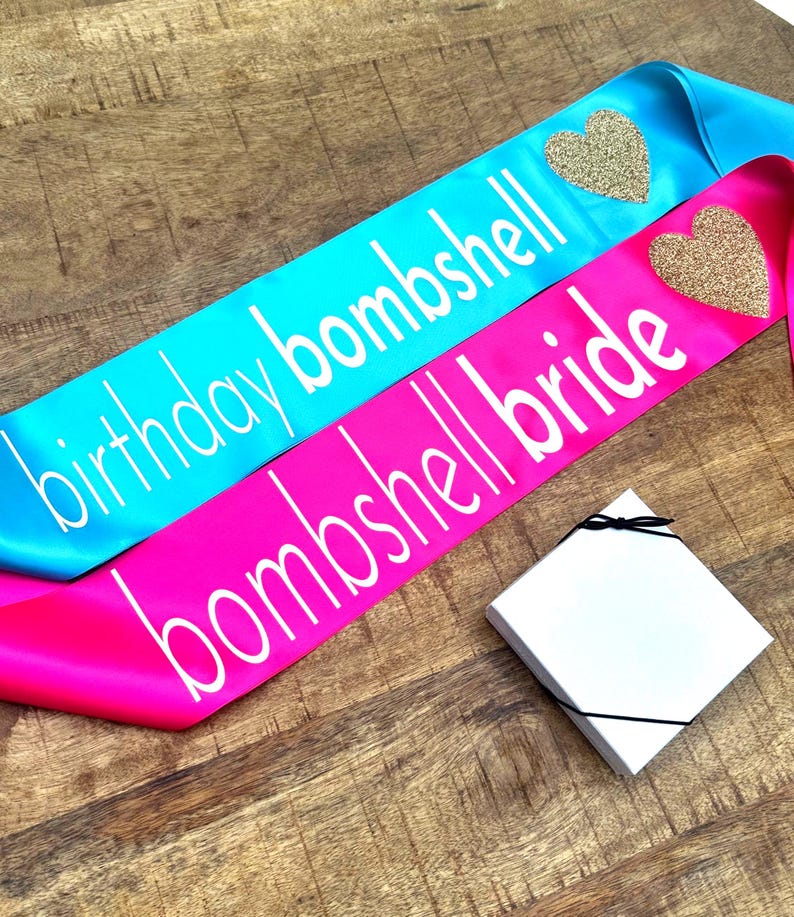 2 sashes the first one read birthday bombshell in white text with glitter gold heart on a turquoise sash.  The second sash reads bombshell bride with a glitter gold heart.  There is also a gift box in the picture with a bow which is included with the sash.