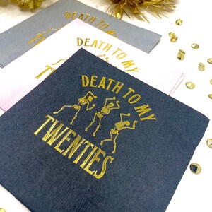 May include: A stack of square cocktail napkins in gray, white, and black. The napkins have gold text that reads "DEATH TO MY TWENTIES" with skeleton illustrations. Gold confetti and tinsel are in the background.