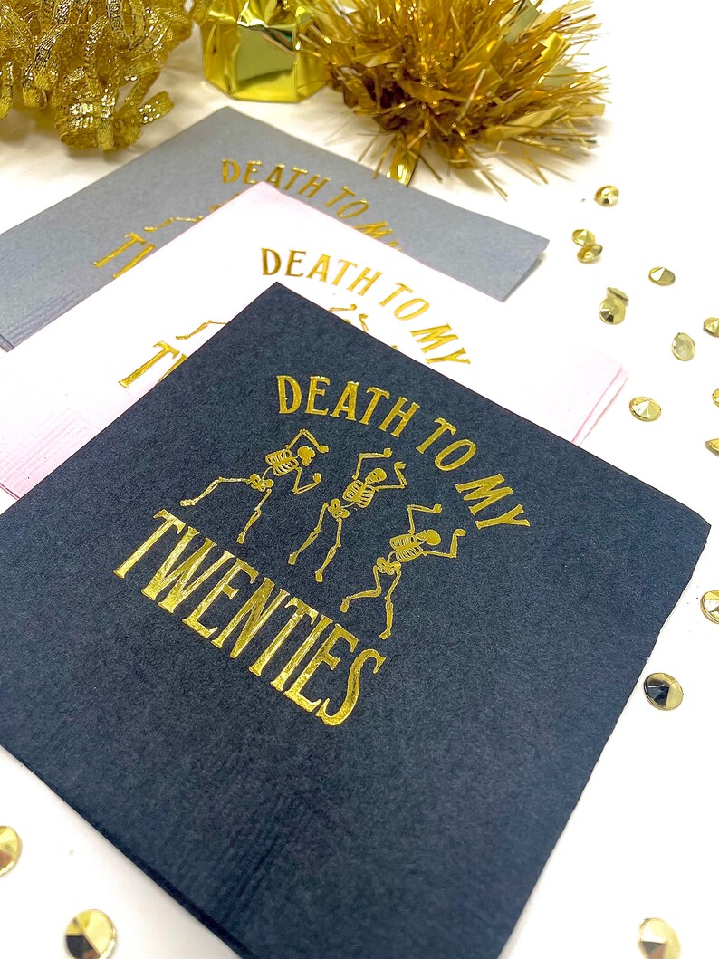 May include: A stack of square cocktail napkins in gray, white, and black. The napkins have gold text that reads "DEATH TO MY TWENTIES" with skeleton illustrations. Gold confetti and tinsel are in the background.