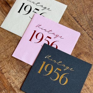 May include: Three square paper napkins with the word "Vintage" and the year "1956" printed on them. One napkin is cream-colored with black text, another is pink with red text, and the third is dark gray with gold text.