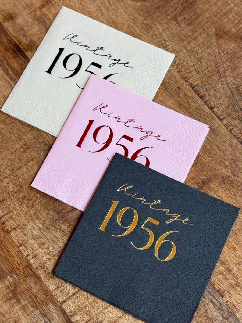 May include: Three square paper napkins with the word "Vintage" and the year "1956" printed on them. One napkin is cream-colored with black text, another is pink with red text, and the third is dark gray with gold text.