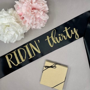 May include: Black satin sash with gold glitter text reading "RIDIN' thirty". The sash is next to a small gift box tied with black string and two large, fluffy paper flowers in white and pink.