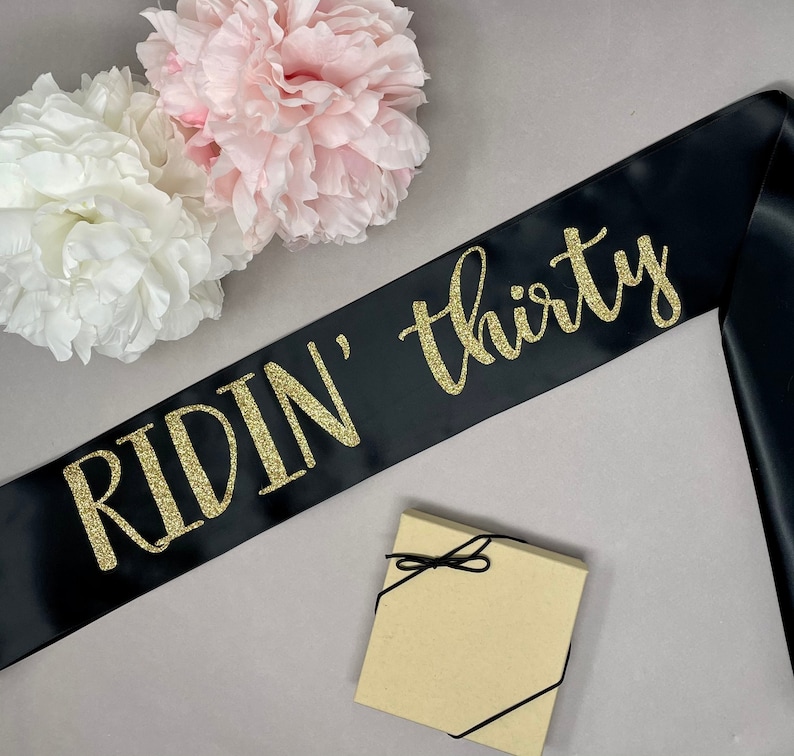 May include: Black satin sash with gold glitter text reading "RIDIN' thirty". The sash is next to a small gift box tied with black string and two large, fluffy paper flowers in white and pink.
