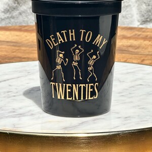 Black plastic cup that reads Death to my twenties in gold text.  With skeletons on the cup.  30th Birthday Plastic Cup