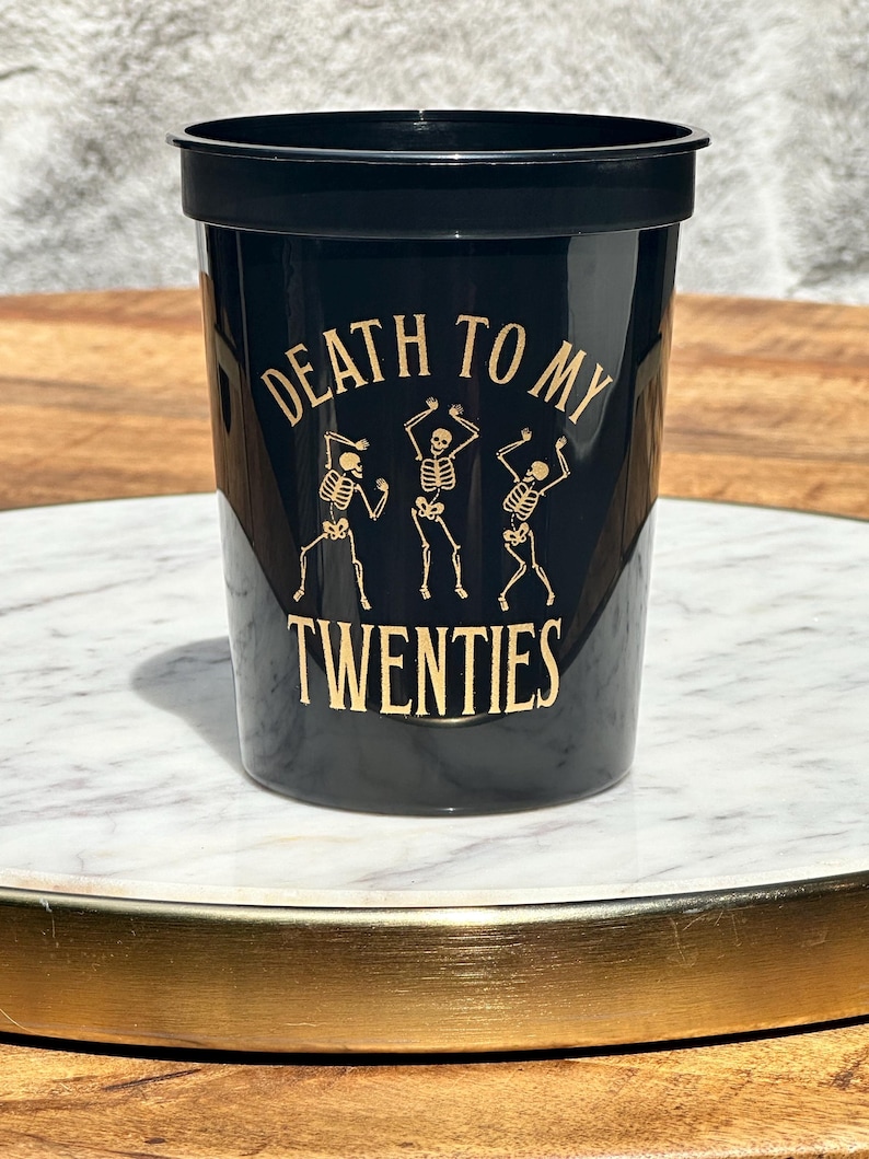 Black plastic cup that reads Death to my twenties in gold text.  With skeletons on the cup.  30th Birthday Plastic Cup