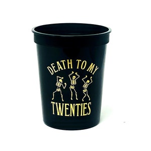 May include: Black plastic cup with gold text and skeleton graphic. The text reads "DEATH TO MY TWENTIES". Three dancing skeletons are featured in the design. The cup is a party or event item.