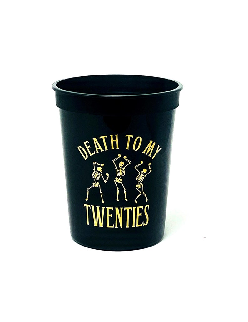 May include: Black plastic cup with gold text and skeleton graphic. The text reads "DEATH TO MY TWENTIES". Three dancing skeletons are featured in the design. The cup is a party or event item.