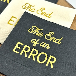 Three square paper napkins, two black and one cream, with "The End of an ERROR" in gold foil script on each.