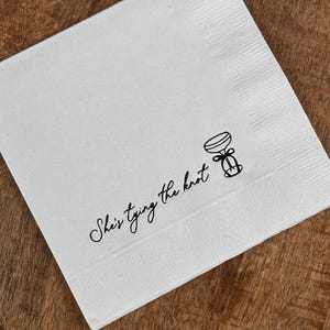 White paper napkin with black text that reads "She's tying the knot" and a simple illustration of a martini with a bow.