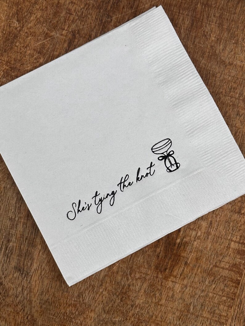White paper napkin with black text that reads "She's tying the knot" and a simple illustration of a martini with a bow.