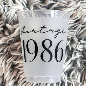 A single frosted plastic cup with "vintage" in black script and "1986" in black block letters is shown on a fuzzy gray background.