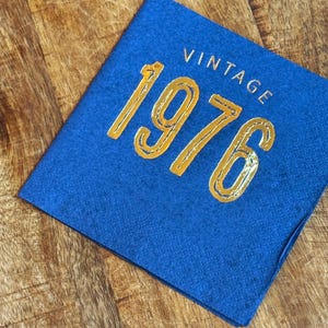 May include: A blue felt square with the word "VINTAGE" above the gold metallic numbers "1976". The item is on a wooden surface.