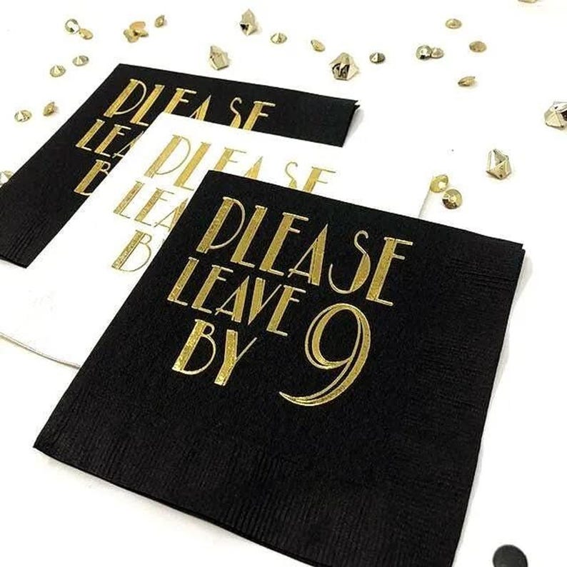 Stacked paper napkins in black and white featuring gold foil stamped text that reads Please Leave By 9, scattered with gold confetti on a white surface.