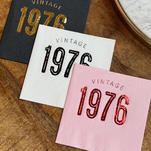 May include: Three square paper napkins with the word "VINTAGE" above the number "1976". One napkin is black with gold lettering, one is white with black lettering, and one is pink with red lettering. The napkins are on a wooden surface.