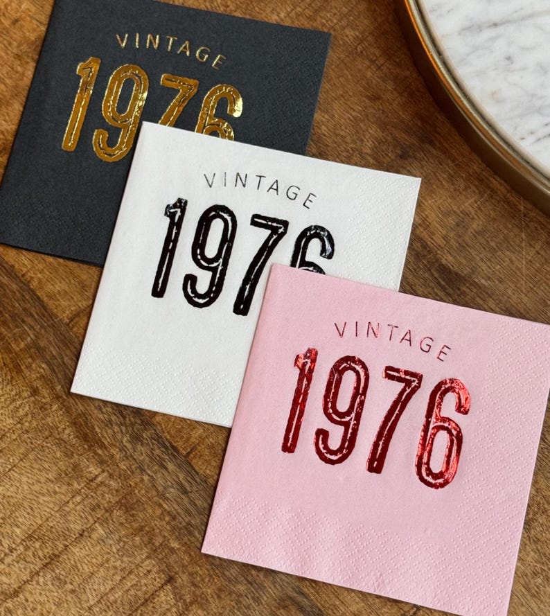 May include: Three square paper napkins with the word "VINTAGE" above the number "1976". One napkin is black with gold lettering, one is white with black lettering, and one is pink with red lettering. The napkins are on a wooden surface.