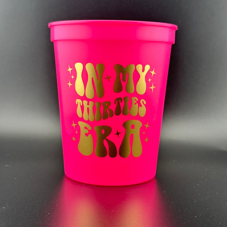 Pode incluir: Um copo de pl&aacute;stico rosa brilhante com um design dourado que diz "In My Thirties Era".
