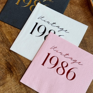 Three square paper napkins are arranged on a dark wooden surface: a dark gray napkin with gold foil text, a white napkin with black foil text, and a pink napkin with red foil text, all reading "Vintage 1986."