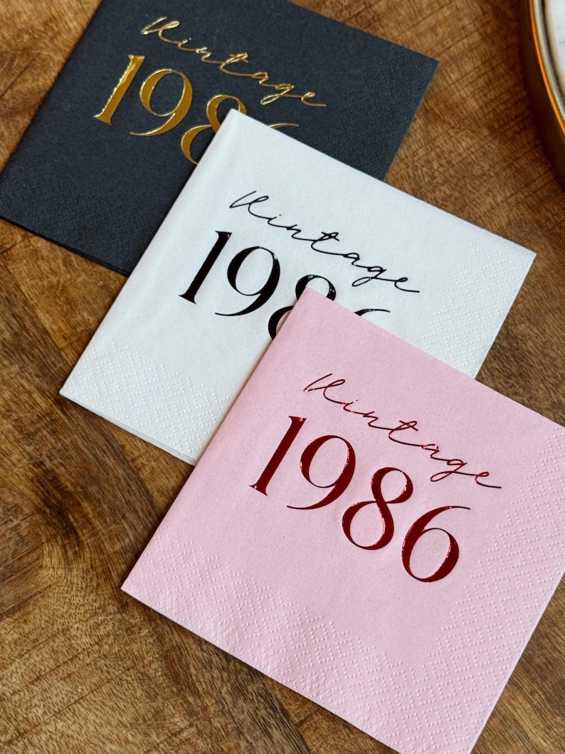 Three square paper napkins are arranged on a dark wooden surface: a dark gray napkin with gold foil text, a white napkin with black foil text, and a pink napkin with red foil text, all reading "Vintage 1986."