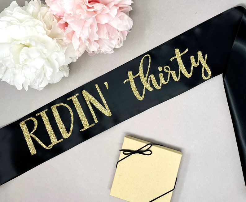Black satin sash with gold glitter text that reads "RIDIN' thirty". The sash is displayed with a small gift box and pink and white flowers in the background. A fun accessory for a milestone birthday celebration.