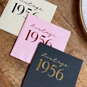 May include: Three square paper napkins in cream, pink, and black, each with the words "Vintage 1956" printed in a script font. The cream napkin has black text, the pink has red, and the black has gold.