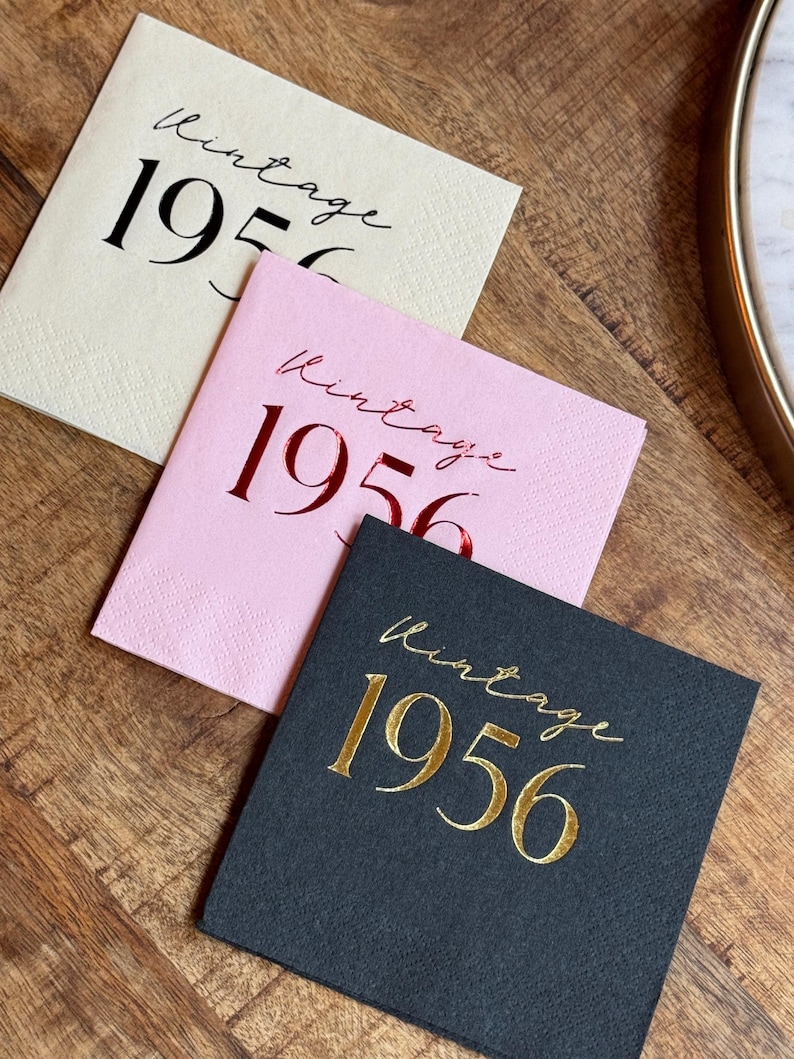 May include: Three square paper napkins in cream, pink, and black, each with the words "Vintage 1956" printed in a script font. The cream napkin has black text, the pink has red, and the black has gold.