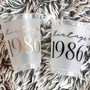 Two frosted plastic cups are shown on a fuzzy gray background, one with gold "vintage 1986" text and the other with black "vintage 1986" text.