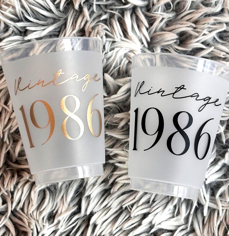 Two frosted plastic cups are shown on a fuzzy gray background, one with gold "vintage 1986" text and the other with black "vintage 1986" text.