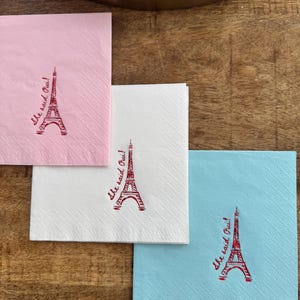 May include: Three square paper napkins in pink, white, and blue, each with a red Eiffel Tower and the phrase "she said Oui!" in red script. The napkins are arranged on a wooden surface, ideal for a celebration.