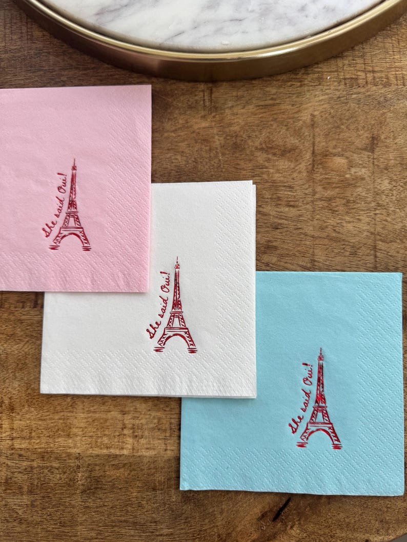 May include: Three square paper napkins in pink, white, and blue, each with a red Eiffel Tower and the phrase "she said Oui!" in red script. The napkins are arranged on a wooden surface, ideal for a celebration.