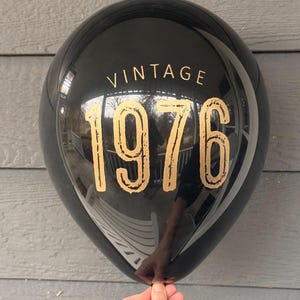 Puede incluir: Globo negro con la palabra "VINTAGE" encima de los n&uacute;meros dorados "1976". El globo est&aacute; sostenido por una mano y refleja el entorno. Es una decoraci&oacute;n de fiesta.