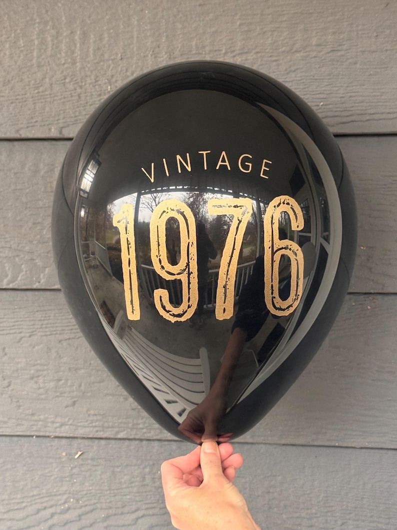 Puede incluir: Globo negro con la palabra "VINTAGE" encima de los n&uacute;meros dorados "1976". El globo est&aacute; sostenido por una mano y refleja el entorno. Es una decoraci&oacute;n de fiesta.