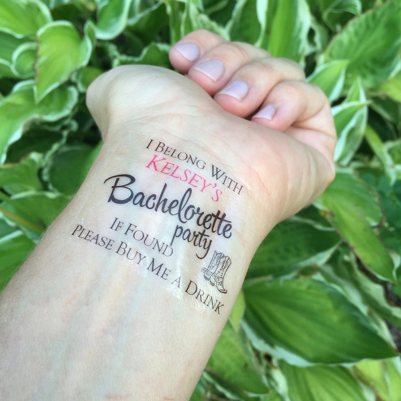 A temporary tattoo on a person's inner wrist reads "I BELONG WITH KELSEY'S Bachelorette party IF FOUND PLEASE BUY ME A DRINK" with two cowboy boots, against a blurred green leafy background.