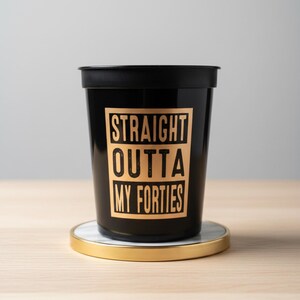 A black plastic cup with the text "STRAIGHT OUTTA MY FORTIES" in a gold-colored font. 50th Birthday Party Favors