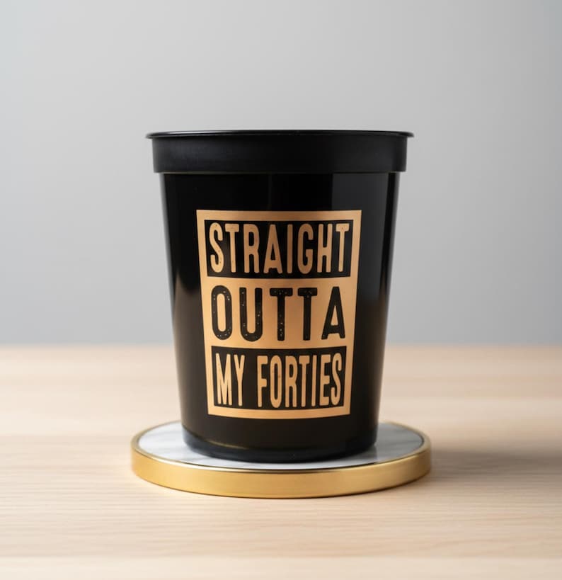 A black plastic cup with the text "STRAIGHT OUTTA MY FORTIES" in a gold-colored font. 50th Birthday Party Favors
