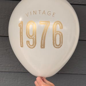 Puede incluir: Un globo blanco con la palabra "VINTAGE" impresa en la parte superior y el n&uacute;mero "1976" en dorado. El globo es sostenido por una mano sobre un fondo gris.