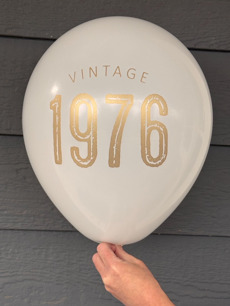 Puede incluir: Un globo blanco con la palabra "VINTAGE" impresa en la parte superior y el n&uacute;mero "1976" en dorado. El globo es sostenido por una mano sobre un fondo gris.