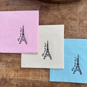 May include: Three square paper napkins in pink, cream, and blue, each with a black Eiffel Tower illustration and the phrase "She said Oui!". The napkins are on a wooden surface.