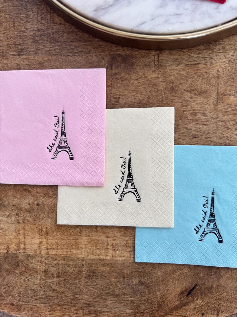 May include: Three square paper napkins in pink, cream, and blue, each with a black Eiffel Tower illustration and the phrase "She said Oui!". The napkins are on a wooden surface.