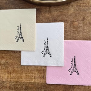 May include: Three square paper napkins in cream, white, and pink, each with a black Eiffel Tower illustration and the script text "She said Oui!". The napkins are on a wooden surface.