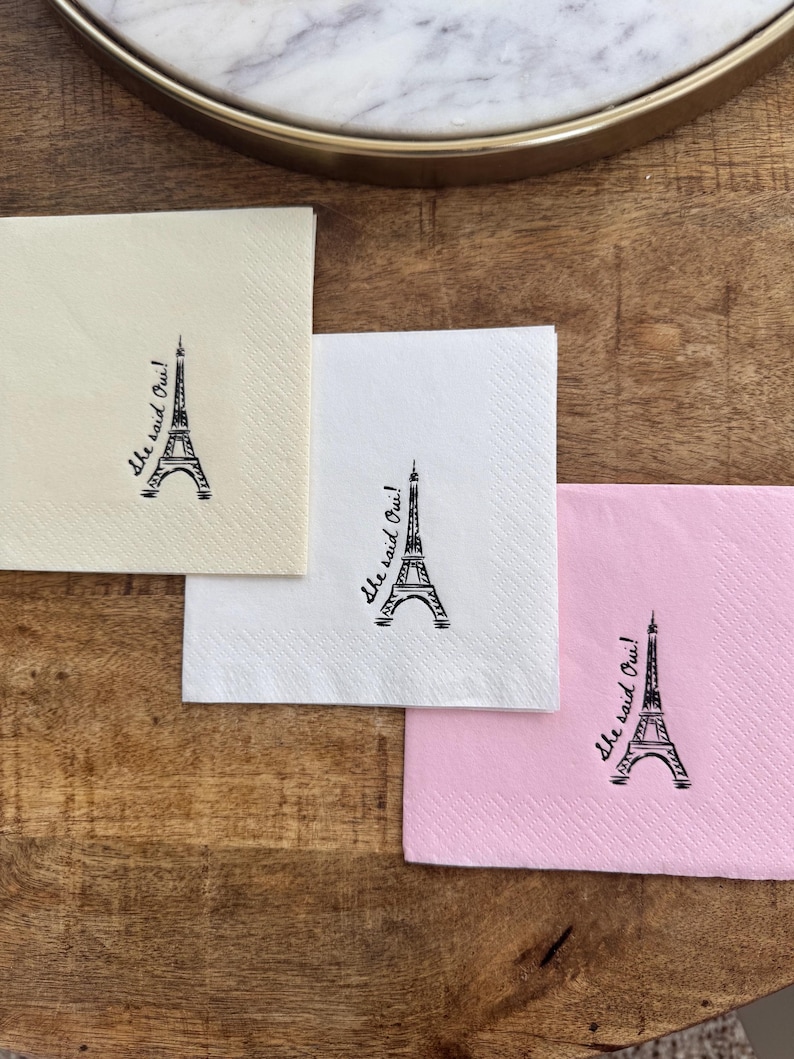 May include: Three square paper napkins in cream, white, and pink, each with a black Eiffel Tower illustration and the script text "She said Oui!". The napkins are on a wooden surface.