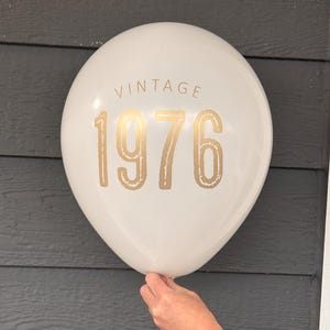 Puede incluir: Globo blanco con texto dorado "VINTAGE 1976". El globo es sostenido por una mano. El fondo es una superficie de madera gris oscuro.