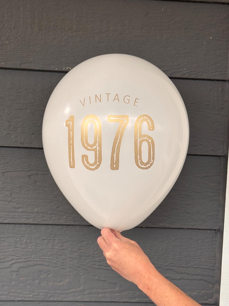 Puede incluir: Globo blanco con texto dorado "VINTAGE 1976". El globo es sostenido por una mano. El fondo es una superficie de madera gris oscuro.