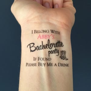 A temporary tattoo on a person's inner wrist reads "I BELONG WITH ABBY'S Bachelorette party IF FOUND PLEASE BUY ME A DRINK" with two cowboy boots, against a dark blue background.
